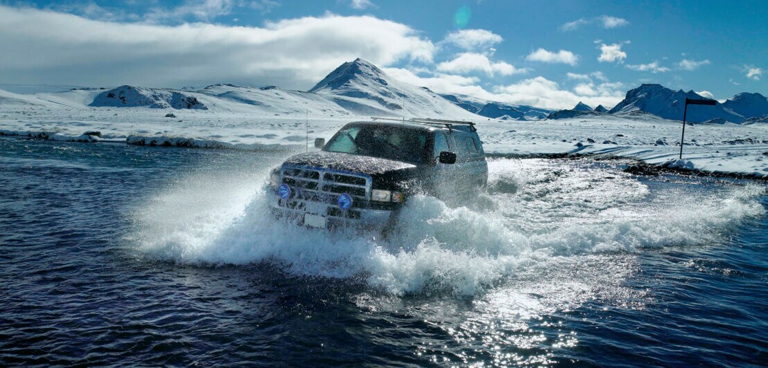 Ein Fahrzeug fährt durch einen Fluss, dahinter erstreckt sich eine Schneelandschaft.