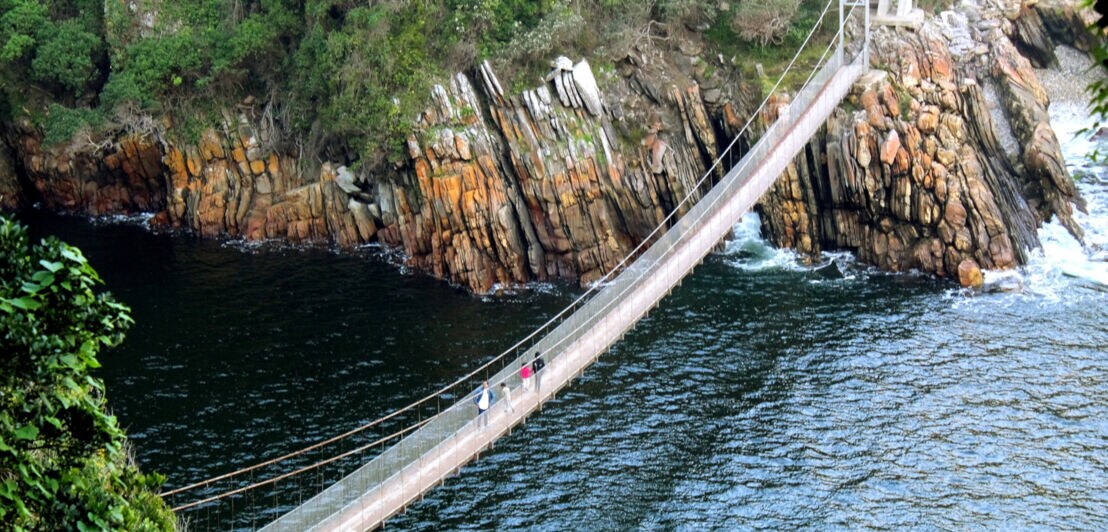Personen auf einer Hängebrücke zwischen bewaldeten Felsklippen über einer Meeresmündung