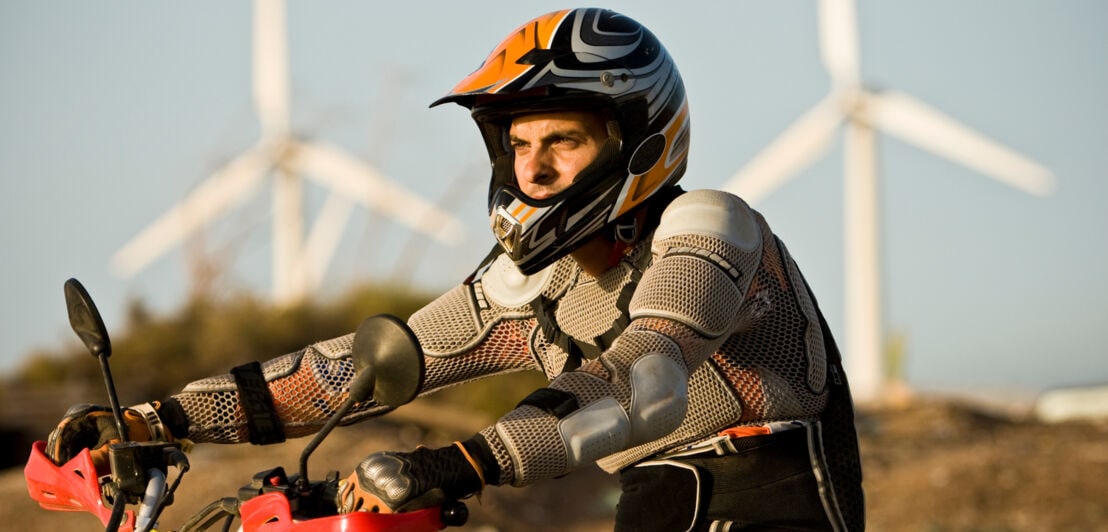Ein Mann in Motocrosskleidung und mit Helm auf einem Bike, im Hintergrund Windräder