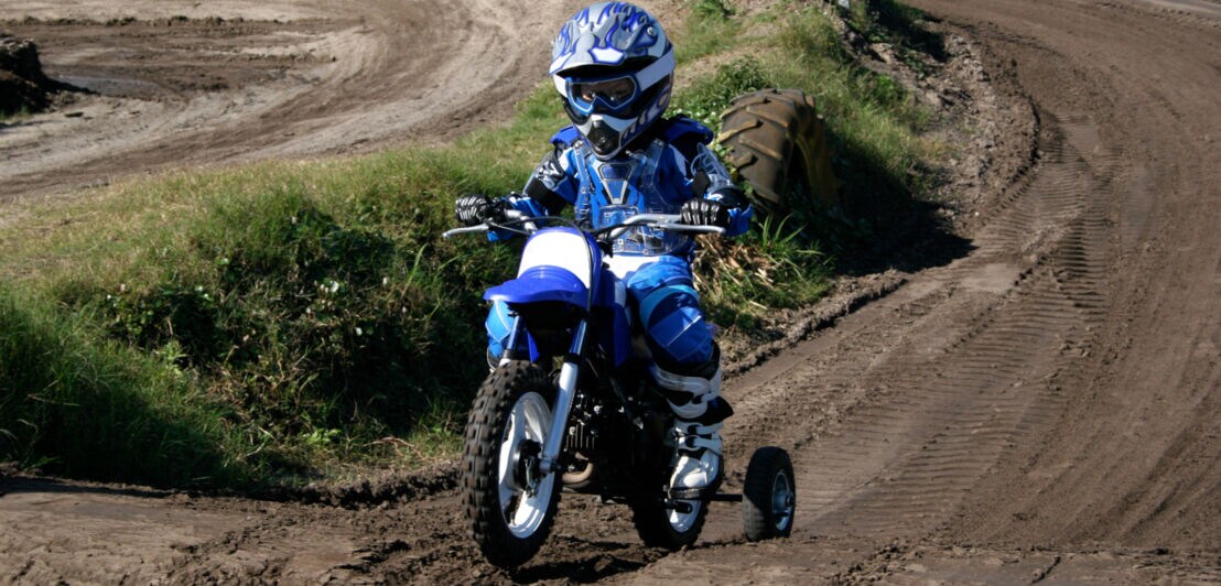 Ein Kind fährt auf einem kleinen Motocross-Bike mit Stützrädern und trägt dabei eine komplette Schutzausrüstung in Blau