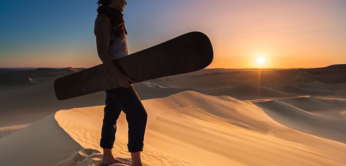 Eine Frau mit einem Sandboard unter dem Arm blickt von einer Sanddüne über eine Wüstenlandschaft in den Sonnenuntergang