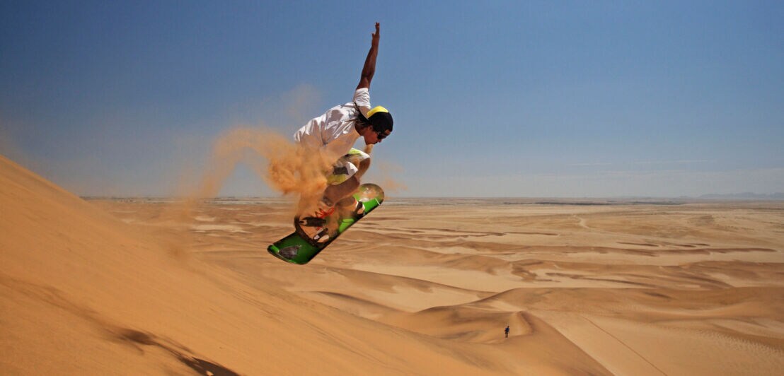 Ein Sandboarder beim Sprung auf einer Sanddüne in einer weiten Wüstenlandschaft