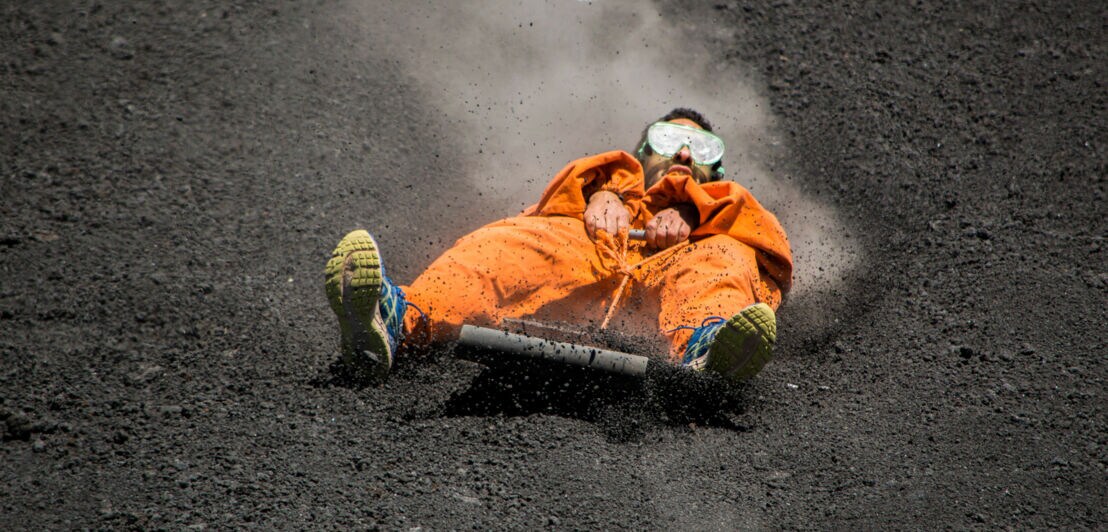 Ein Mann in einem orangefarbenen Schutzanzug fährt auf einem Board liegend über schwarze Vulkanasche einen Abhang hinunter