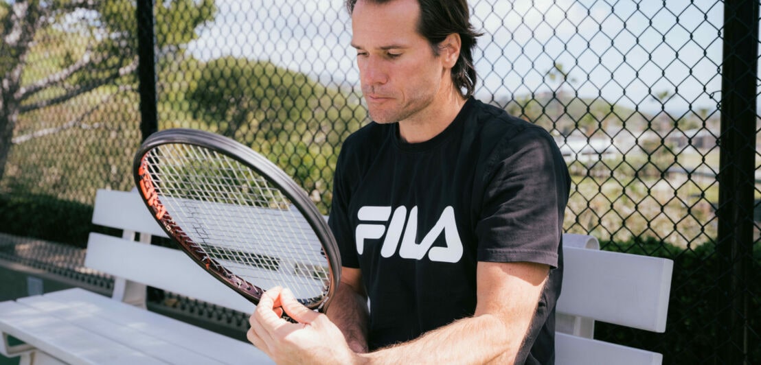 Tommy Haas sitzt auf einer Bank und zupft an den Saiten seines Tennisschlägers.