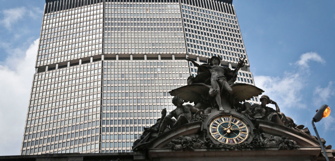 Das geradlinige MetLife-Hochhaus erhebt sich gen Himmel, im Vordergrund eine verspielte Skulptur mit Uhr am Eingang eines Gebäudes im Beaux-Arts-Stil