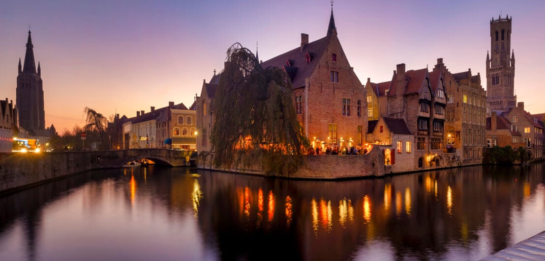 Personen an einer beleuchteten Kaimauer am Wasserkanal in der mittelalterlichen Altstadt von Brügge bei Abenddämmerung.