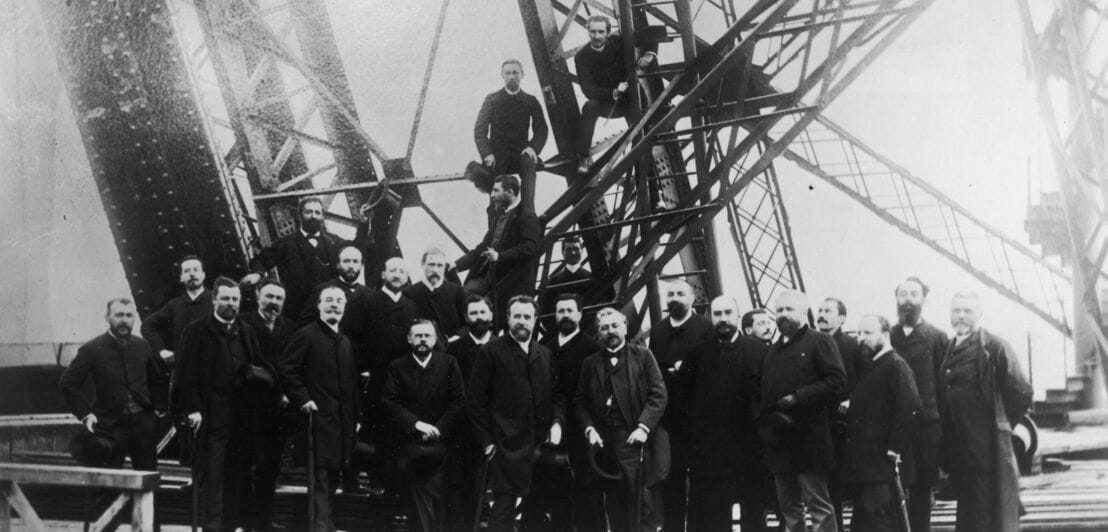 Schwarzweißaufnahme von Gustave Eiffel mit einer Gruppe Männer vor einer Stahlkonstruktion beim Bau des Eiffelturms.