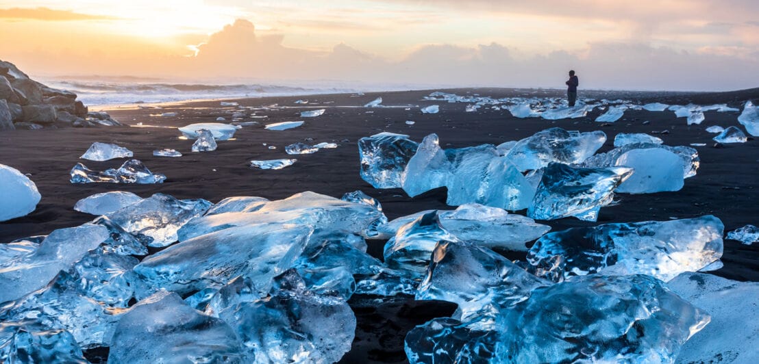 Eine Person steht am Diamond Beach in Island und blickt auf blaue Eisbrocken, die auf dem schwarzen Sand liegen und im Licht der aufgehenden Sonne glitzern