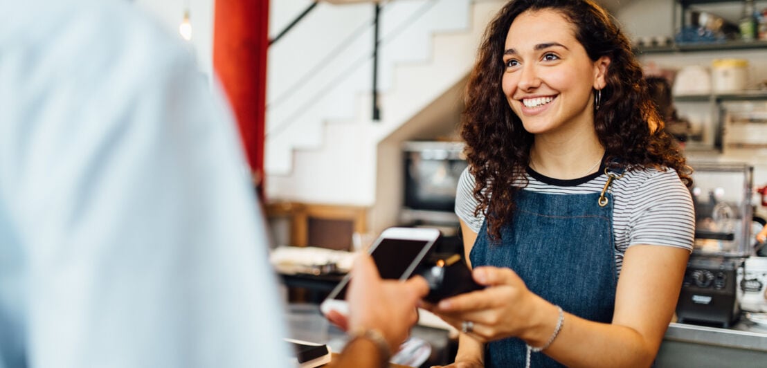 Ein Kunde bezahlt via Mobile Payment bei einer lächelnden Verkäuferin am Tresen in einem Café