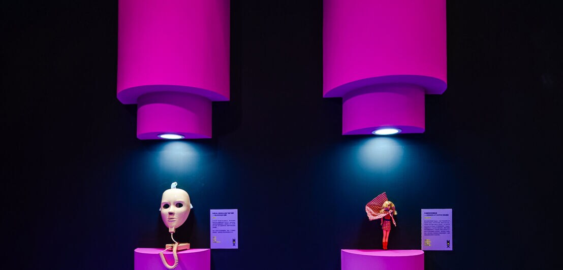 Eine Gesichtsmaske aus Plastik und eine Puppe als Ausstellungsexponate an einer Wand.