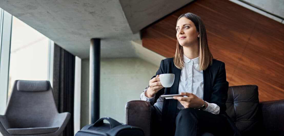 Eine junge Geschäftsfrau genießt die Wartezeit mit einer Tasse Kaffee auf einem Sessel in einer Lounge