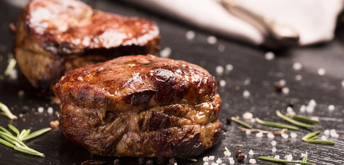 Gegrilltes Steakfleisch, grobes Meersalz und Rosmarinnadeln auf einer dunklen Oberfläche