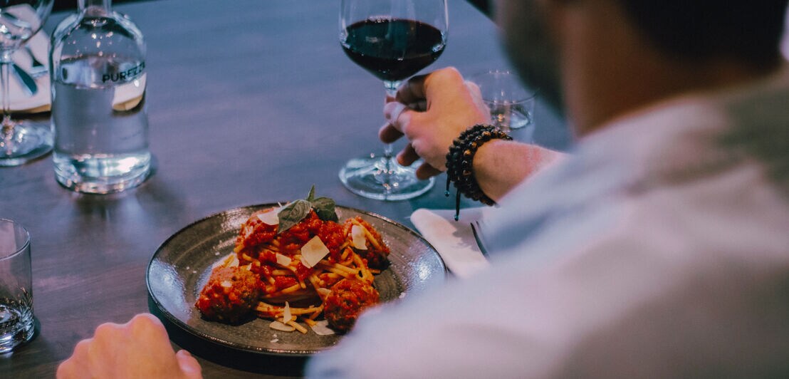 Blick über die Schulter eines Mannes an einem Restauranttisch, der vor sich einen Teller mit Essen und ein Glas Wein hat