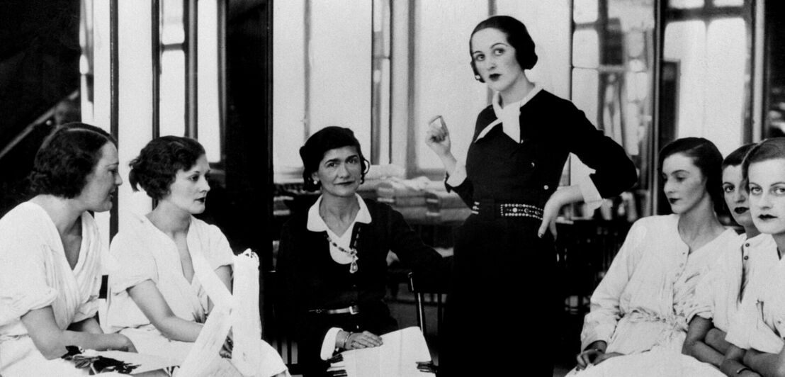 Schwarzweißfoto von Coco Chanel in einem schwarzen Kostüm, umgeben von weiblichen Models in ihrem Modeatelier