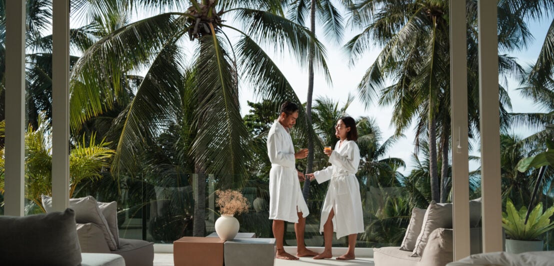 Ein Paar steht in weißen Bademänteln auf der Terrasse eines Luxushotels in tropischer Umgebung