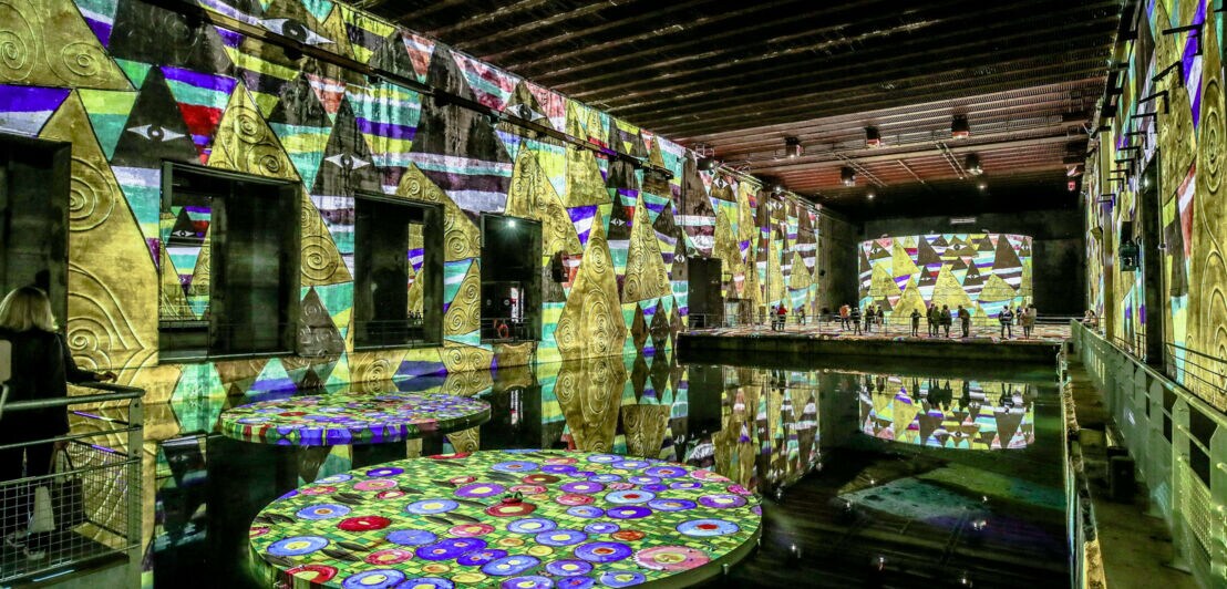 Besucher einer digitalen Lichtausstellung in einer alten Fabrikhalle mit an die Wände projizierten Kunstwerken des Malers Gustav Klimt