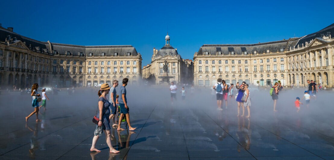 Personen in Sommerbekleidung laufen über einen Wasserspiegel auf einem großen Platz vor neoklassizistischen Gebäuden in Bordeaux