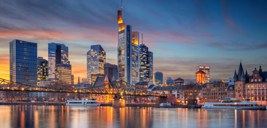 Erleuchtete Skyline von Frankfurt hinter einer Brücke über einen Fluss mit Booten bei Abenddämmerung