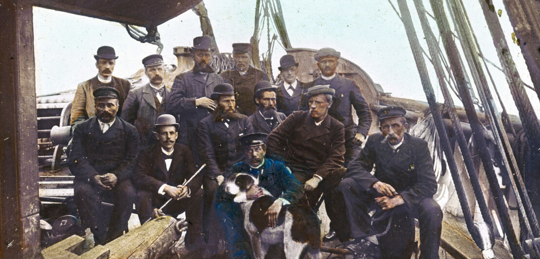 Eine Gruppe von Männern in Arbeitsanzügen und mit Hüten sowie ein Hund an Bord eines Schiffes 