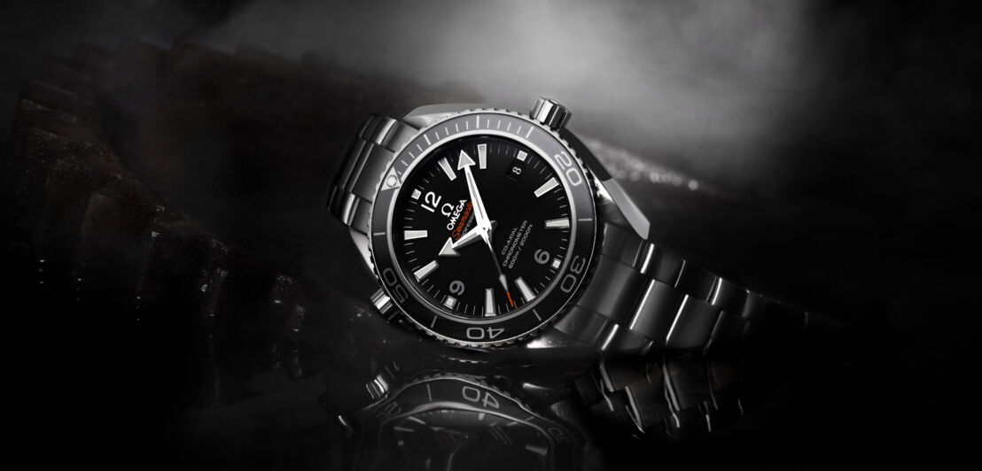 Produktaufnahme, Close-up einer liegenden OMEGA Uhr mit schwarzem Zifferblatt und metallenem Gliederarmband auf schwarzem Untergrund