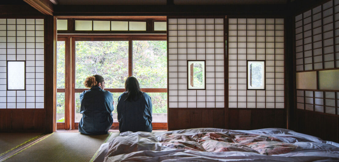 Rückansicht zweier Personen, die neben einem Futonbett in einer japanischen Herberge sitzen und aus dem Fenster in einen Garten schauen