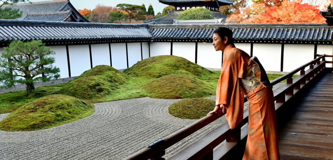 Eine Geisha in einem orangefarbenen Kimono blickt von einer Terrasse in einen Zen-Garten im Zentrum einer Tempelanlage