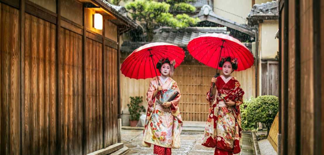 Zwei junge Geishas in rot-weiß-gemusterten Kimonos und weiß geschminkten Gesichtern laufen mit roten Regenschirmen durch eine Gasse in Kyoto