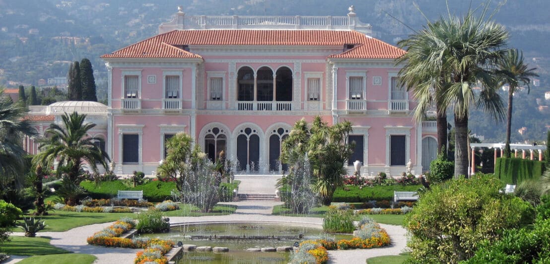 Blick auf die rosafarbene Fassade der Villa Ephrussi de Rothschild, vor der sich ein Teich erstreckt und die in einer gepflegten Parkanlage steht