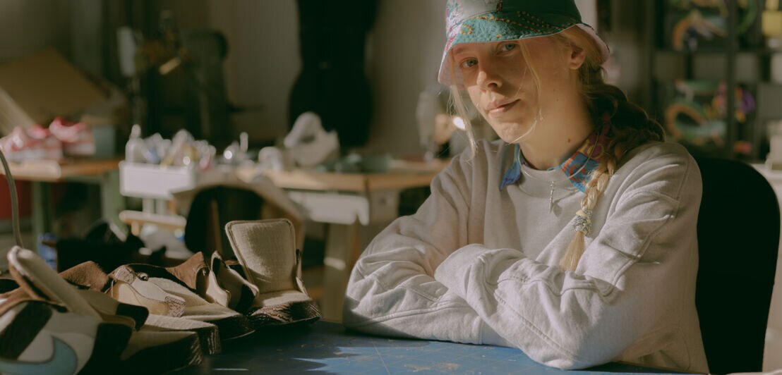 Eine Frau mit Hut und blondem Zopf sitzt an einem Arbeitstisch, auf dem Turnschuhe stehen