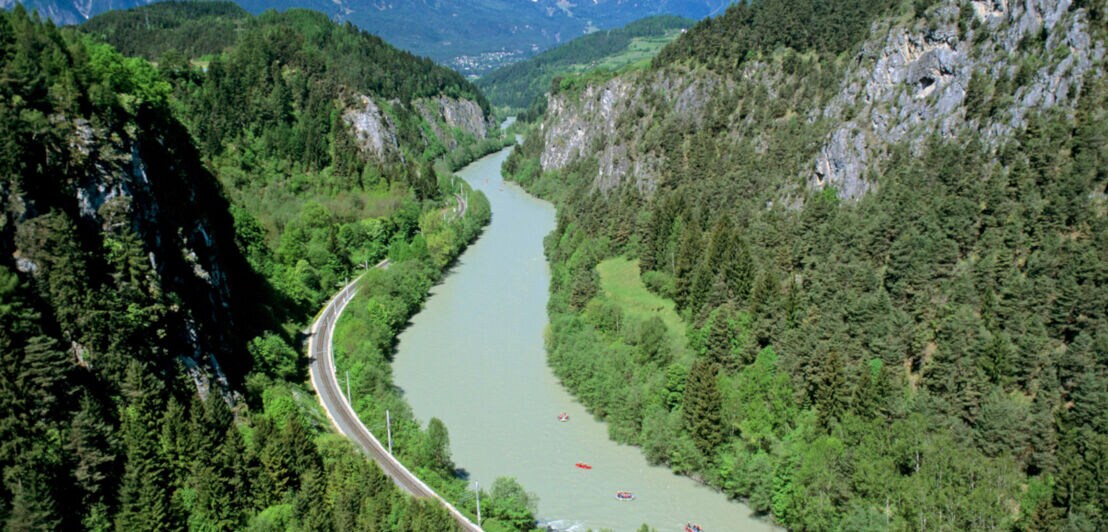 Alpenpanorama mit Waldgebiet und einem breiten Fluss, auf dem Raftingboote fahren