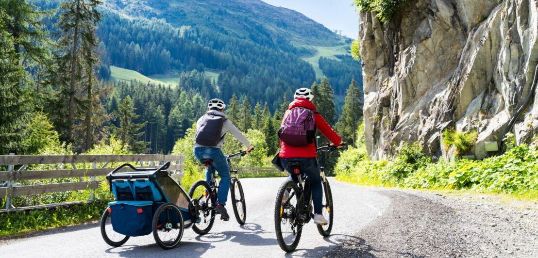 Rückansicht eines Paares mit Rucksäcken auf E-Bikes, das auf einer Straße durch eine Berglandschaft fährt; an einem Fahrrad befindet sich ein Kinderanhänger