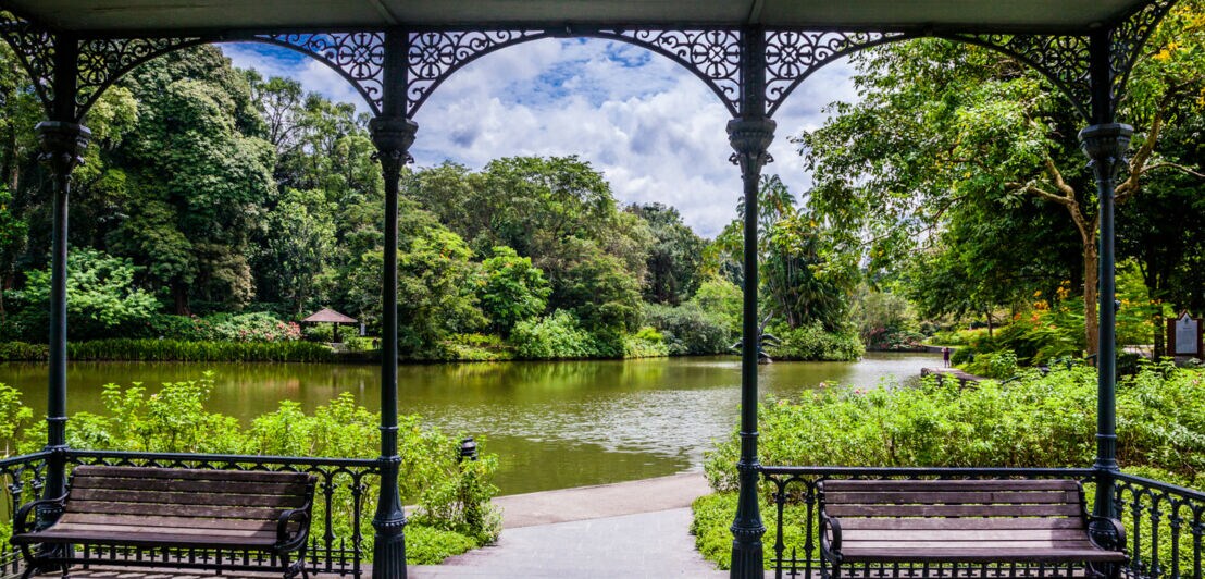 Blick auf einen See in einer grünen Parkanlage aus einem Pavillon mit Holzbänken