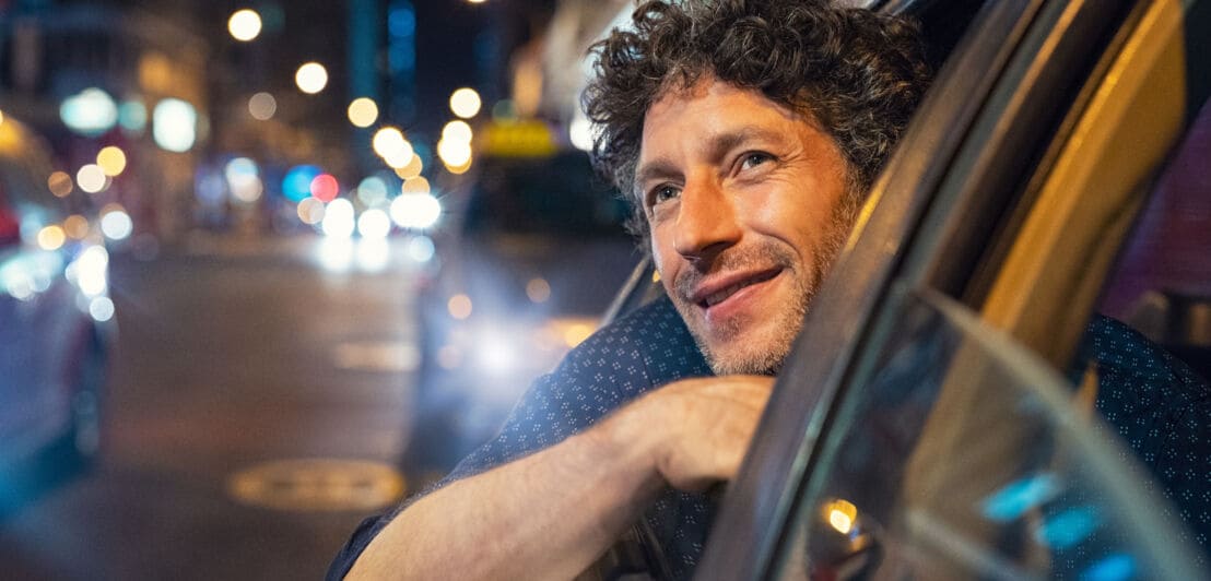 Ein Mann auf dem Rücksitz eines Autos blickt lächelnd aus dem Fenster eines fahrenden Autos im Stadtzentrum.