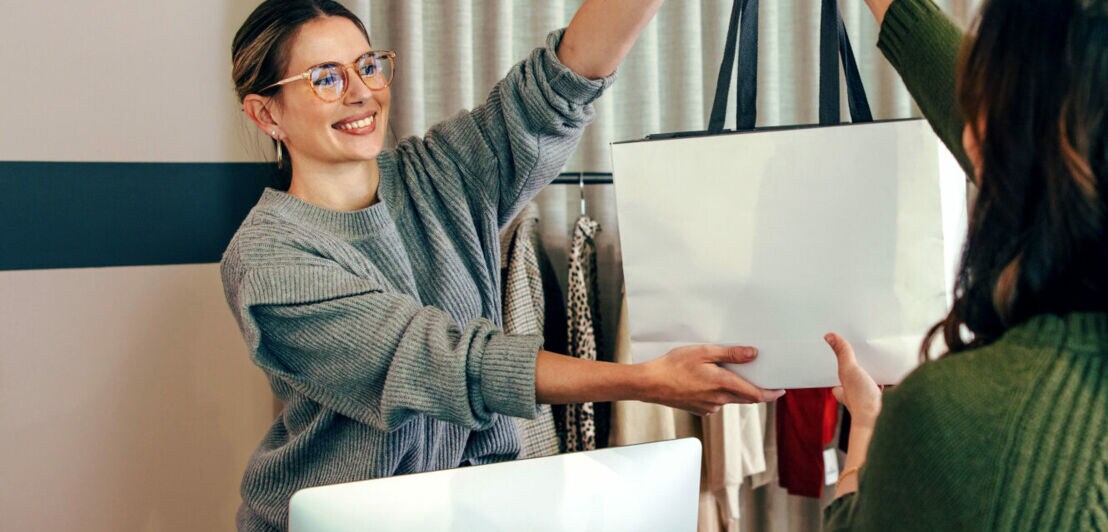 Eine lächelnde Verkäuferin reicht einer Kundin eine Einkaufstasche über den Kassentresen in einer schicken Modeboutique