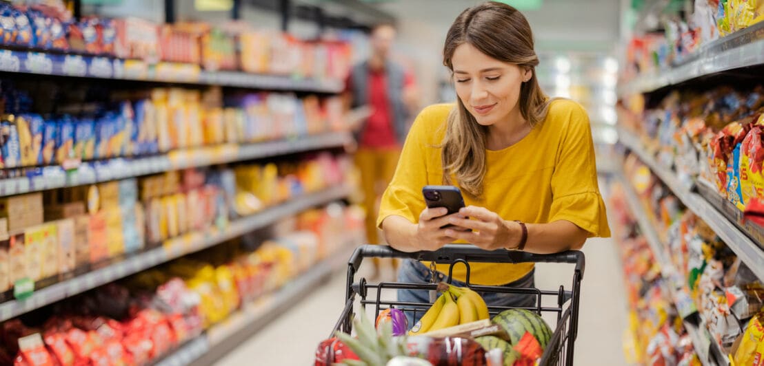 Eine junge Frau im Supermarkt lehnt an ihrem vollgepackten Einkaufswagen und schaut auf ihr Smartphone