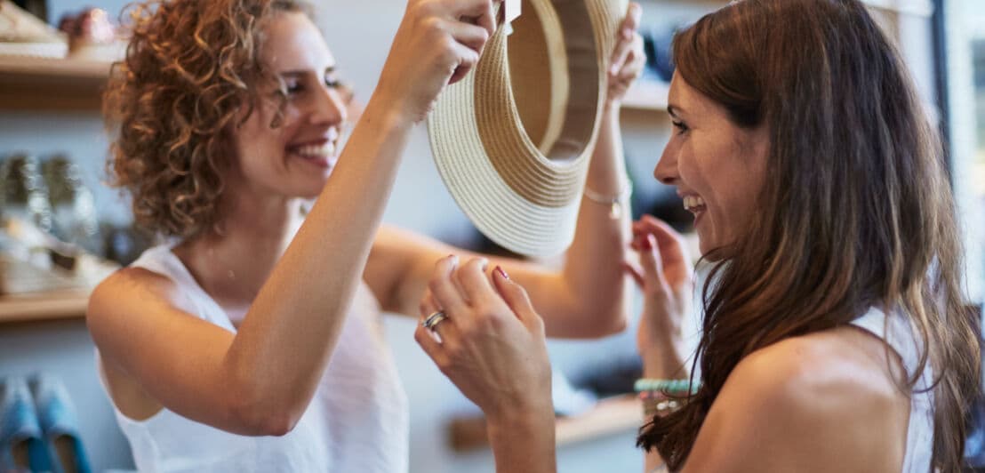 Zwei Freudinnen amüsieren sich beim Anprobieren eines Hutes in einer Modeboutique