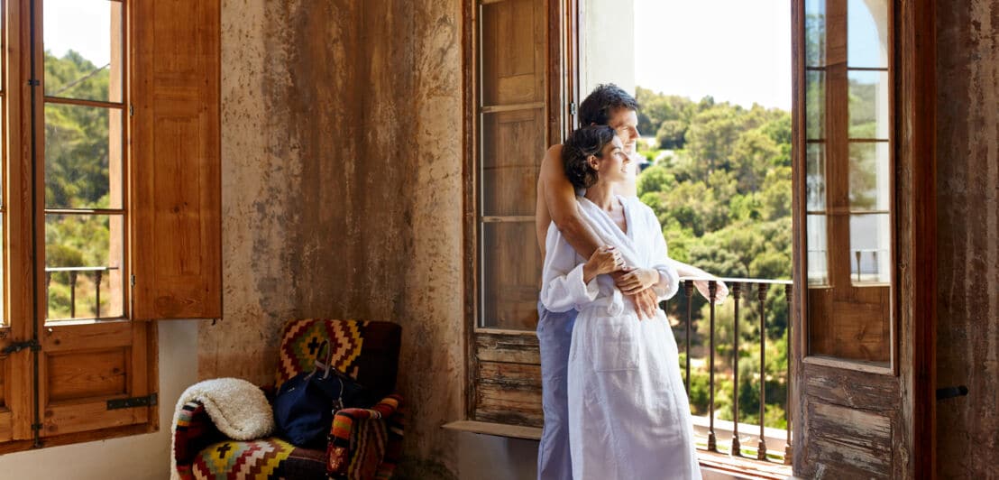 Ein sich umarmendes Paar steht an einem großen Fenster in einer mediterranen Hotelsuite und schaut in die grüne Landschaft; sie trägt einen weißen Bademantel