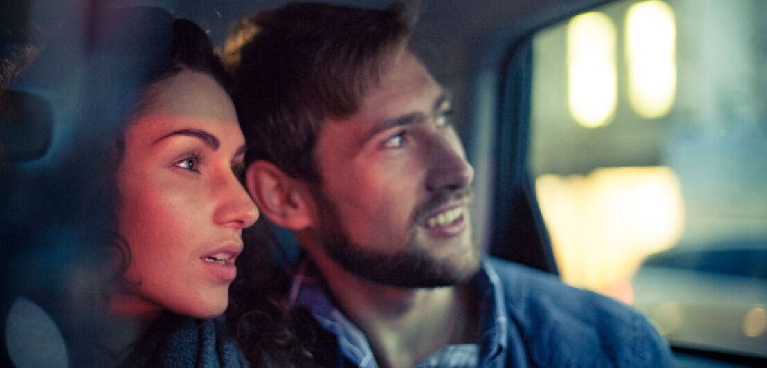 Nahaufnahme eines Paares, das, angeleuchtet von den Lichtern einer Stadt, staunend aus einem Autofenster blickt.