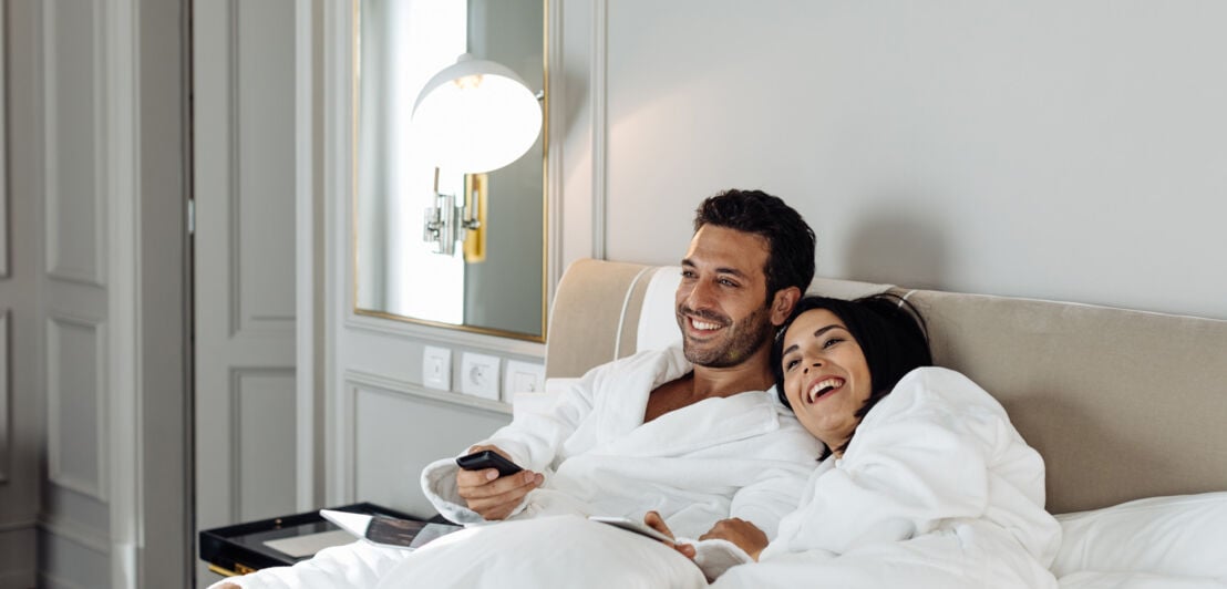 Ein fröhliches Paar in weißen Bademänteln entspannt im Bett einer edlen Hotelsuite