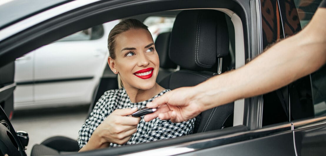 Eine junge Frau sitzt am Steuer eines Mietwagens und nimmt lächelnd den Autoschlüssel durch das geöffnete Fenster entgegen