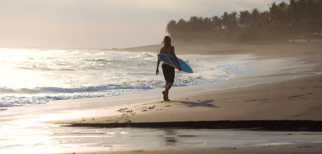 Rückansicht einer Person mit Surfbrett unter dem Arm, die einen palmengesäumten Sandstrand in der Abendsonne entlangläuft