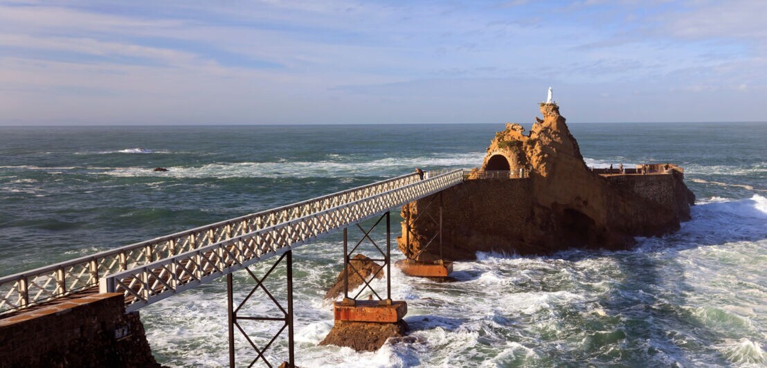 Eine Eisenbrücke führt zu einem Felsen im Meer, auf dem eine Marienstatue steht