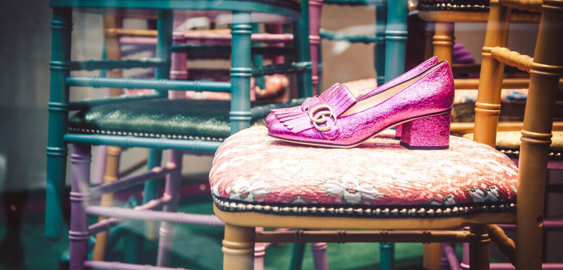 Ein Paar glitzernde, pinkfarbene Loafer mit goldenem Gucci-Logo steht auf einem Stuhl in einem Schaufenster