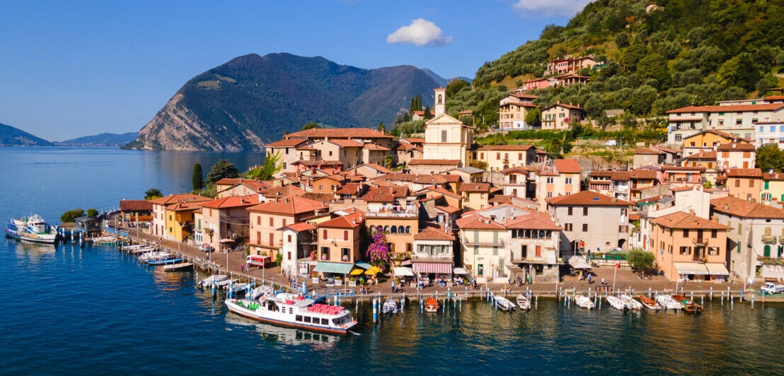 Kleiner Küstenort am Iseosee mit Promenade und Booten im Wasser am Fuße eines bewaldeten Berges