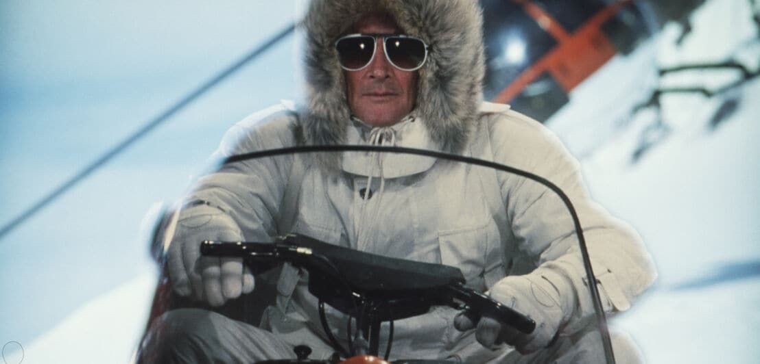 Schauspieler Roger Moore fährt im weißen Schneeanzug und großer Sonnenbrille auf einem Schneemobil
