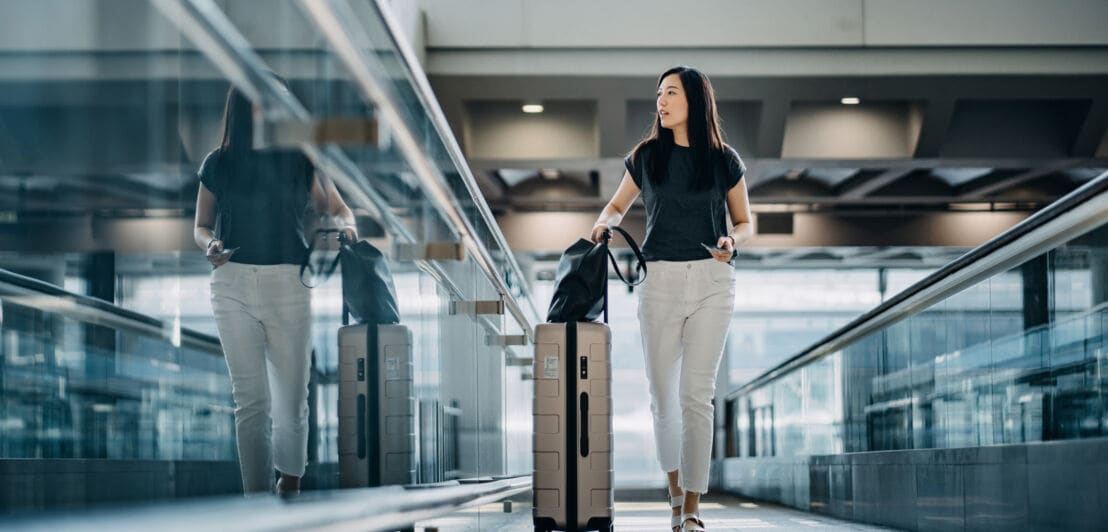 Eine elegante, junge Frau läuft mit einem Rollkoffer durch eine Eingangshalle am Flughafen