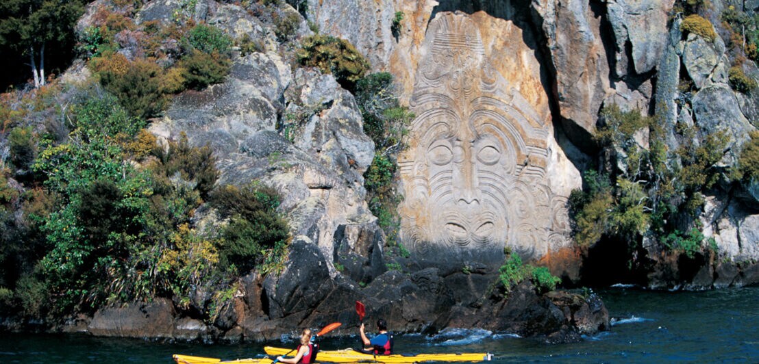 Zwei Kajakfahrer paddeln auf einem Gewässer entlang einer Felswand mit Maori-Kunst