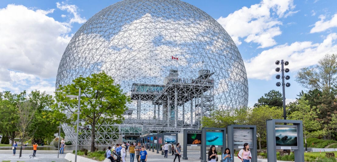 Die Stahlkuppel Biosphère in Montreal, umrahmt von Bäumen, davor Menschen