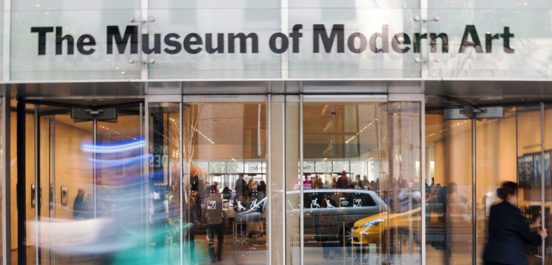 Personen am Eingang des Museum of Modern Art mit Glastüren, in denen sich auf der Straße vorbeifahrende Autos spiegeln.
