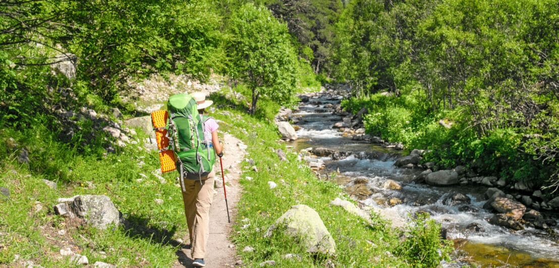 Rückansicht einer Wanderin mit Rucksack und Wanderstock, die auf einem schmalen Weg entlang eines Flusses durch eine grüne Naturlandschaft mit Bergpanorama läuft